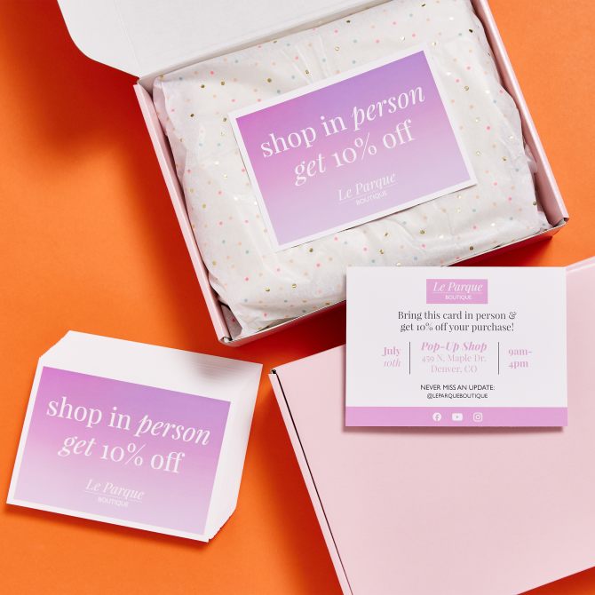 A promotional setup featuring postcards for a boutique business. The front of the card reads "shop in person get 10% off" in white text on a purple gradient background. The back of the card provides details for a pop-up shop event. The cards are displayed on top of a white box lined with polka dot tissue paper and an orange background. The brand's social media handles are included at the bottom of the card.