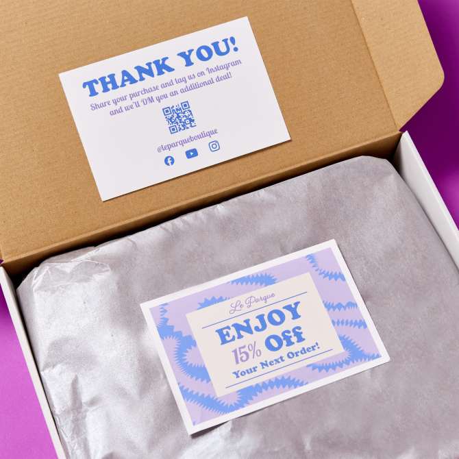 An example of a packaging insert made with a double-sided postcard. One side of the postcard reads "ENJOY 15% Off Your Next Order!" in blue text on a cream and purple background with a starburst design. The other side displays a thank you message: "THANK YOU! Share your purchase and tag us on Instagram and we'll DM you an additional deal!" along with the store's social media handles and a QR code. The postcard is displayed on silver tissue paper inside a cardboard box with a purple background.