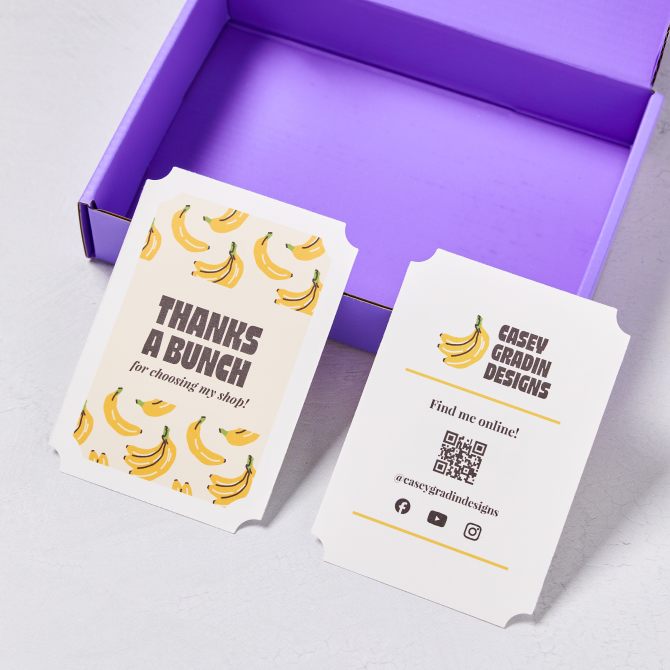 An example of a promotional double-sided 3-1/2" x 5" cards. One side of the card has a playful design with yellow bananas and the text "THANKS A BUNCH for choosing my shop!" in bold letters. The other side features the brand's name, a banana graphic, a QR code, and social media handles to connect with the store. The cards are displayed on a light surface next to an open purple box.