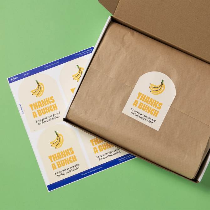 An open box with brown paper lining and a label for product packaging that says 'THANKS A BUNCH' with an illustration of bananas. Next to the box is a sheet of identical labels with the same design and the phrase 'Keep your eyes peeled for fun stuff inside!