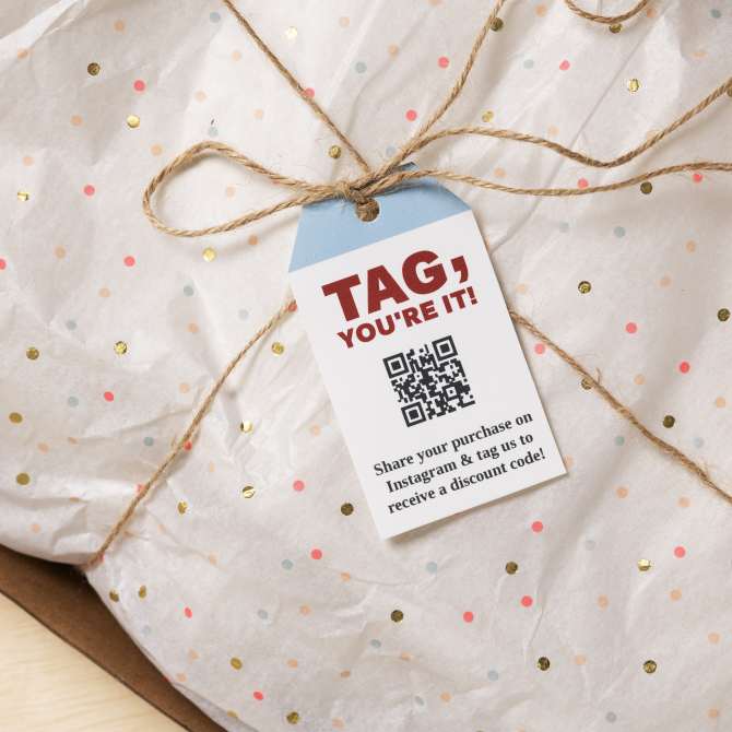 A package wrapped in white tissue paper with colorful dots, tied with twine. Attached to the twine is a tag that reads 'TAG, YOU'RE IT!' with a QR code below it. The text at the bottom of the tag says, 'Share your purchase on Instagram & tag us to receive a discount code!