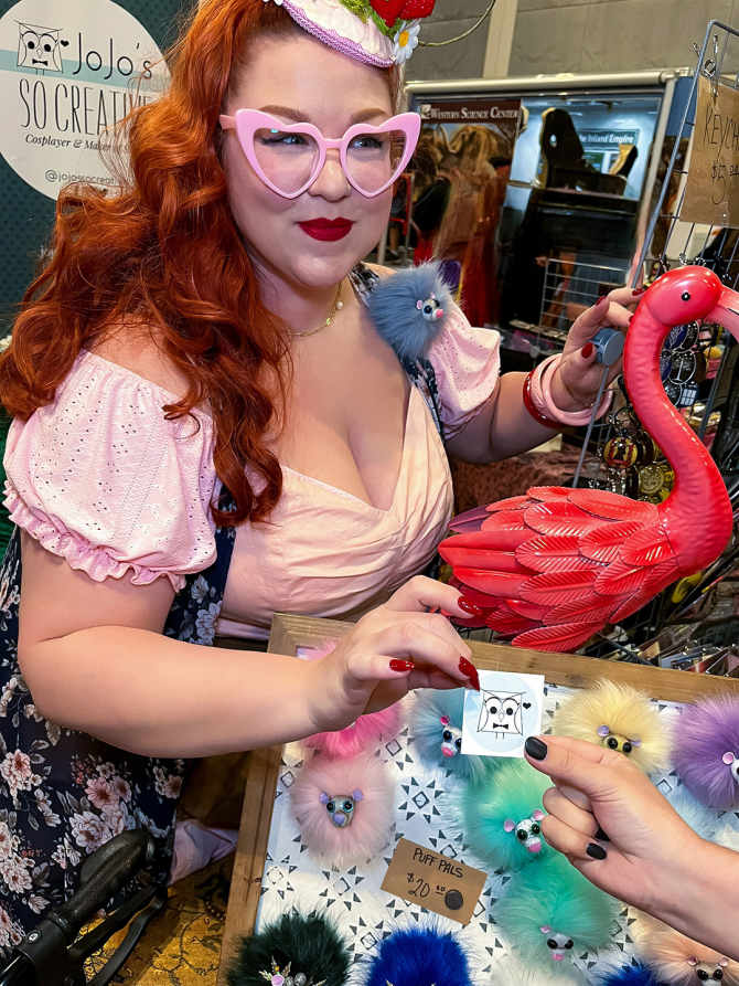 A redheaded woman with pink heart shaped glasses and wearing a pink cottage core dress hands a free sticker to an attendee at Ontario Comic Con Revolution from inside her vendor booth. 