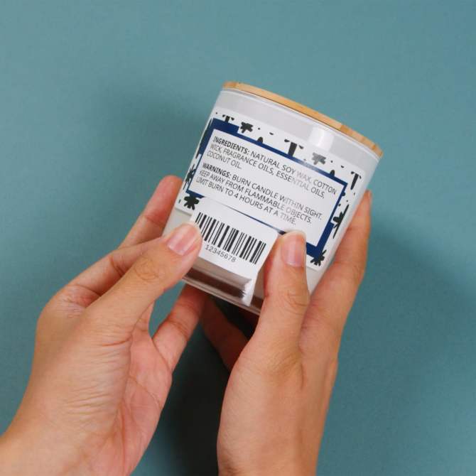 Hands holding a candle jar with an Avery label featuring TrueBlock technology applied to cover previous text and barcodes. The label includes product ingredients and warnings, showing how Avery’s TrueBlock labels effectively hide old information for a clean, new look.