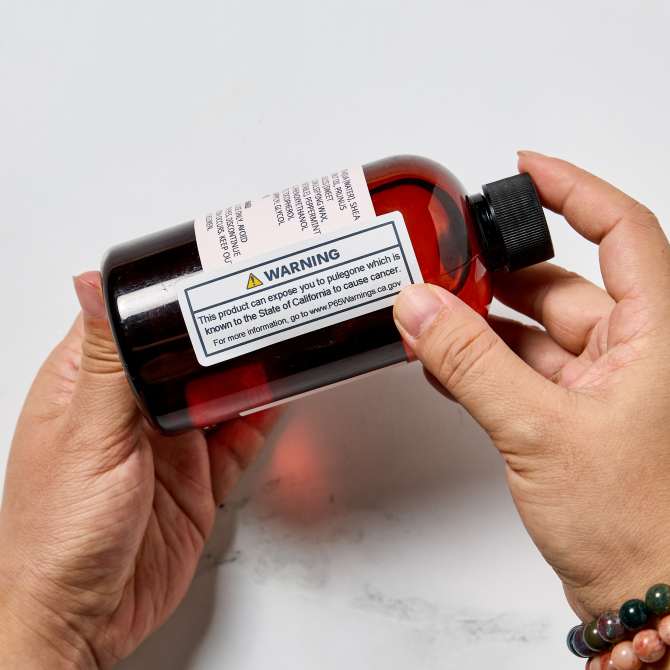 Hands applying an Avery label with TrueBlock technology to a dark amber bottle. The label displays a product safety warning, demonstrating how Avery’s TrueBlock labels fully cover existing labels, ensuring that old information is completely hidden for clear and professional labeling.