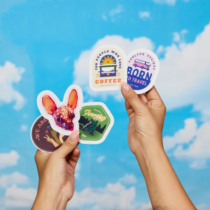 Two hands hold up a variety of colorful stickers made by Avery, a Sticker Mule alternative, against a bright blue sky with fluffy white clouds. The stickers feature different designs, including a dog illustration, a coffee-themed sticker, a travel-themed sticker, a forestry-themed sticker, and one with the phrase "We are." The stickers showcase vibrant colors and distinct shapes, standing out clearly against the sky.