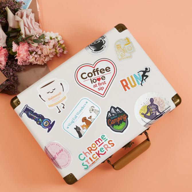 A white suitcase decorated with a variety of durable stickers made by Avery, a Sticker Mule alternative. The stickers include designs such as a heart-shaped "Coffee love at first sip," a "RUN" graphic, a camping-themed sticker, and a colorful "Chrome Stickers" label. The suitcase sits on a peach-colored surface next to a bouquet of flowers, showcasing the vibrant, high-quality stickers that are perfect for personalizing travel gear.