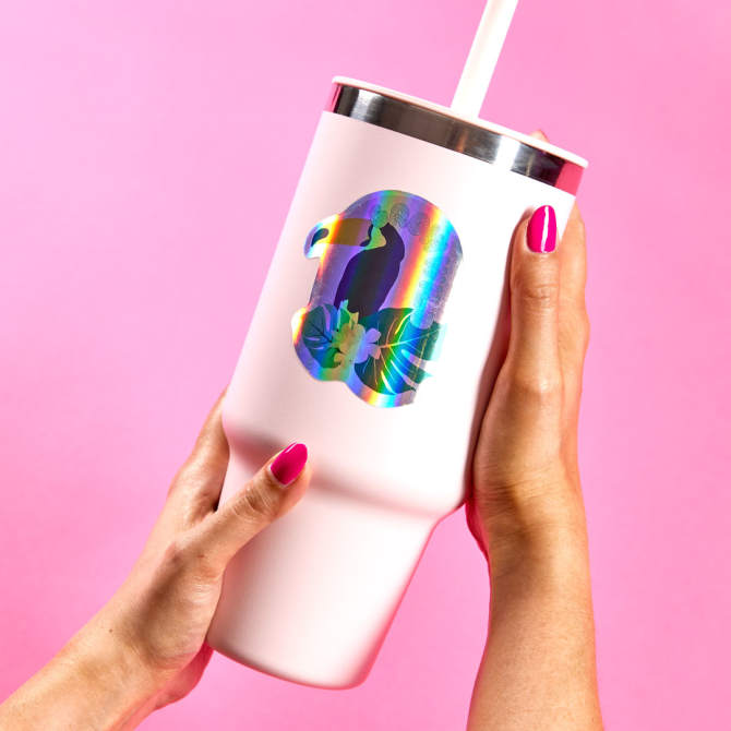 A person holds a white tumbler adorned with a vibrant holographic sticker featuring a tropical bird and floral design. The sticker, made by Avery, stands out with its iridescent colors against the matte finish of the tumbler. The scene is set against a pink background, highlighting the premium quality and eye-catching effect of the Avery sticker, a Sticker Mule alternative.