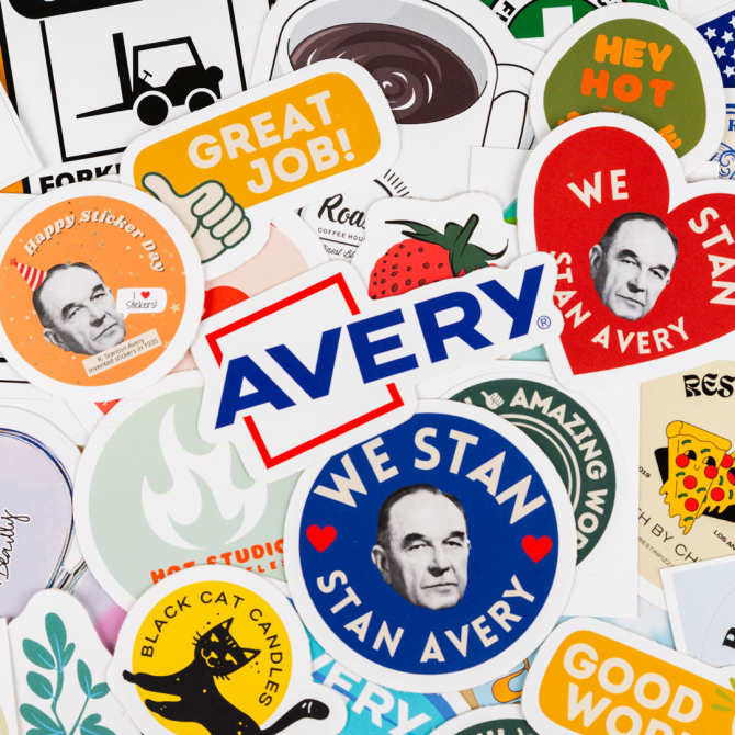 A close-up view of a vibrant assortment of stickers made by Avery, a Sticker Mule alternative. The stickers include various designs, such as "We Stan Stan Avery" featuring an image of Stan Avery, the person that invented stickers and founded the original Avery company, a bold "AVERY" logo, and other playful graphics like a coffee cup, a thumbs-up with "Great Job!", and a strawberry. The stickers are arranged in a colorful, overlapping pattern, highlighting the variety and quality of Avery's sticker products.