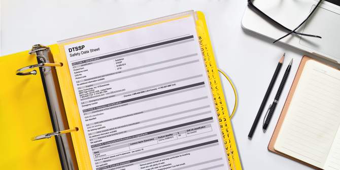 Alt text: A safety data sheet (SDS) is displayed in a yellow Avery SDS binder with Avery alphabetical dividers visible on the right side. The SDS includes sections such as "Identification" and "Hazards Identification," essential for workplace safety and compliance. The setup, complete with pencils, a pen, glasses, and a notebook, suggests a well-organized and prepared workspace.