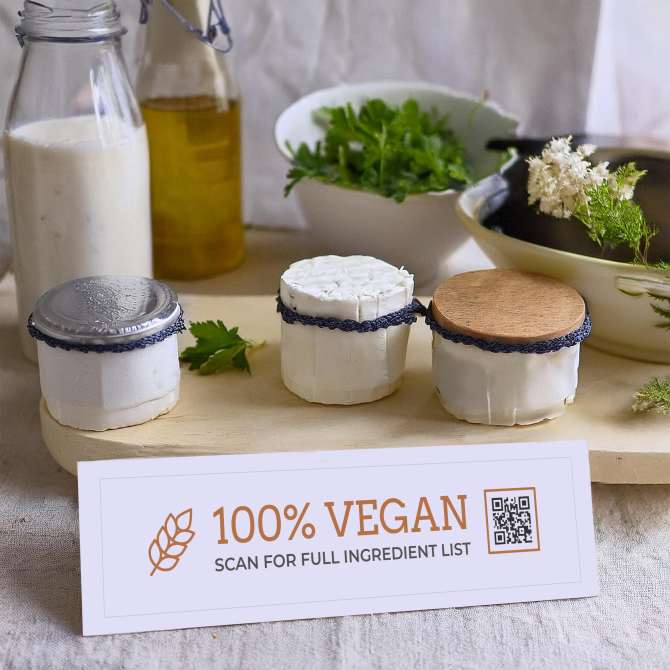 Image showing a farmers market display of vegan cheeses wrapped in paper and twine with herbs and bottles of oil in the background. In front, an Avery tent card reads "100% Vegan" with a QR code that invites customers to "Scan for full ingredient list."