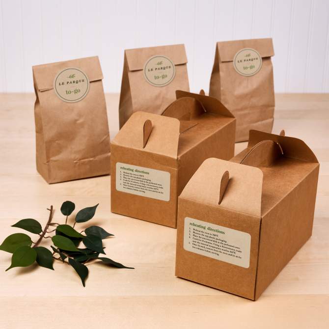  Image of a collection of kraft paper to-go bags and boxes, labeled with custom Avery stickers featuring reheating directions and the branding. The display highlights eco-friendly packaging options for farmers market or food business stands.