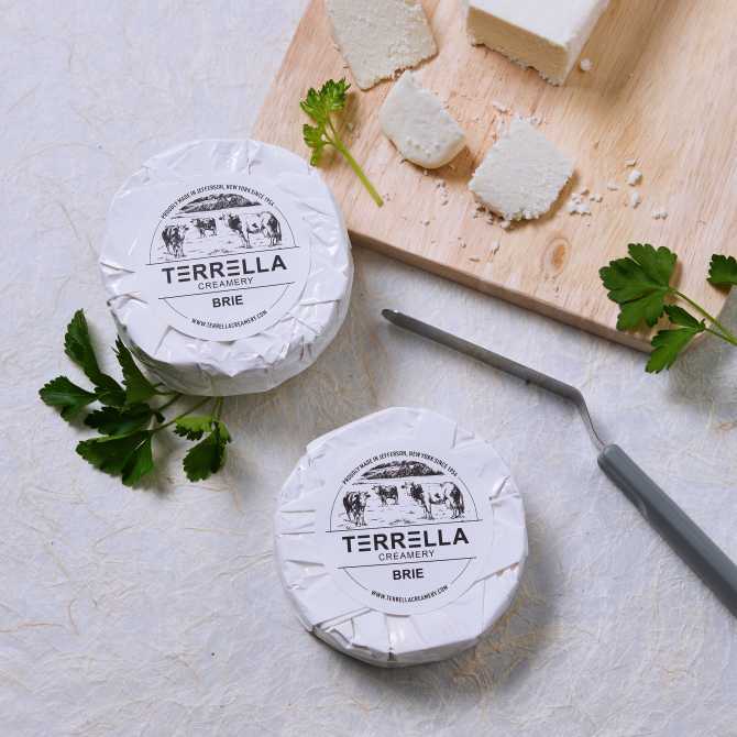 Image showing two rounds of cheese wrapped in paper with a round Avery label featuring a custom farmers market label template with a black and white logo of cows. The cheese is displayed on a light background, alongside a cutting board with cheese slices and a cheese knife.