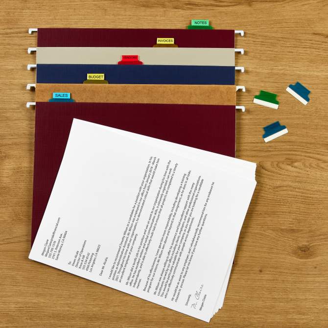 Avery hanging file folders on a wooden desk, each labeled with colorful tabs for categories such as "Notes," "Invoices," "Vendor," "Budget," and "Sales." A stack of documents sits nearby, with additional tab inserts shown beside the folders. This setup demonstrates the use of Avery printable hanging file folder labels for efficient organization.