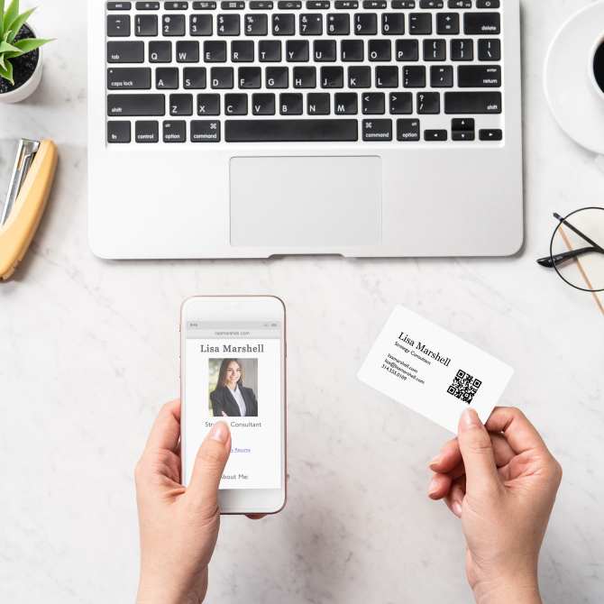 A person holds a business card with a QR code generated using the Avery barcode generator, alongside a smartphone displaying an online profile. The card, designed for "Lisa Marshell, Strategy Consultant," includes contact details and the scannable QR code, which links directly to her website. The setup is placed on a white marble desk with a laptop, a pair of glasses, a small plant, and office supplies, emphasizing a professional, tech-savvy workspace.