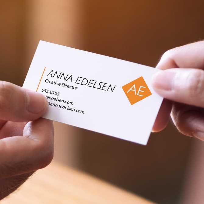 A close-up of a person holding a business card created with Avery Business Card product 28877. The card features a modern layout with the name "Anna Edelsen," title "Creative Director," and contact information on a clean, white background. A stylized "AE" logo in orange appears on the right side, adding a pop of color and a professional touch.