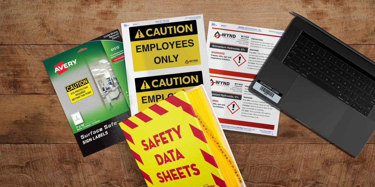 A top-down view of a rugged wooden work table with Avery safety supplies arranged around a laptop. The Avery products include printable safety signs and labels showing caution and GHS chemical safety messages as well as a bright yellow and red Safety Data Sheet binder with dividers.