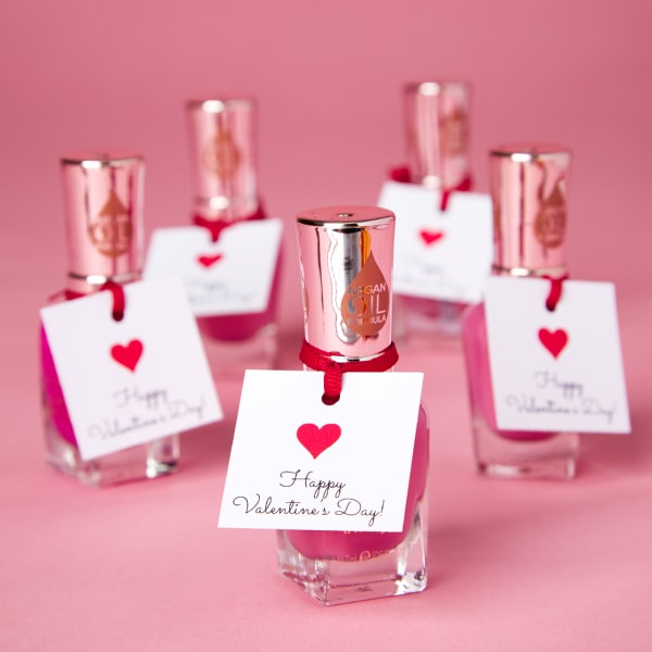 five small square tags with happy Valentine's Day message tied to five pink nail polishes with red string, sitting on pink table