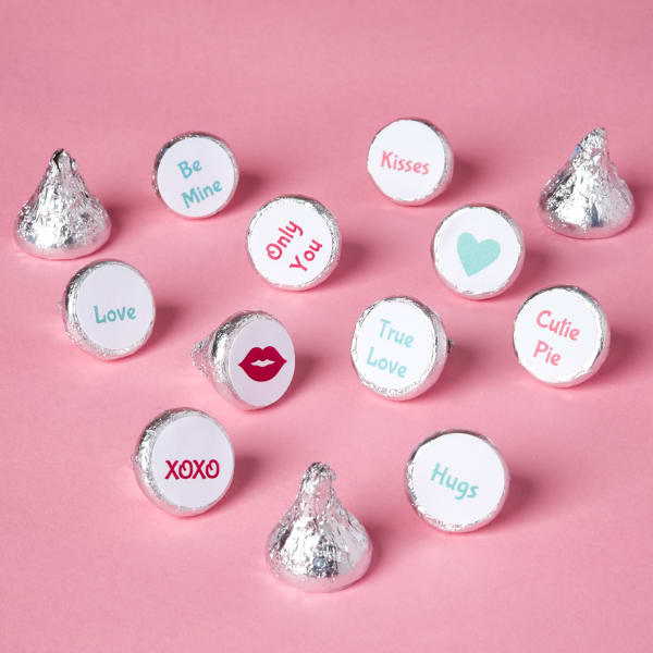 several Hershey's chocolate kisses in silver wrapping labeled with tiny, round Valentine's Day themed messages and illustrations on the bottoms, sitting on pink table