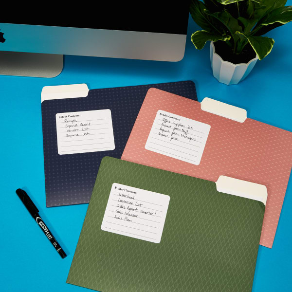 three file folders — one in dark blue, one in blush pink, and one in sage green laying flat on bright blue desk next to Mac