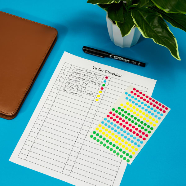 Small color coding label sheet next to a to do checklist with labels on bright blue desk next to a plant