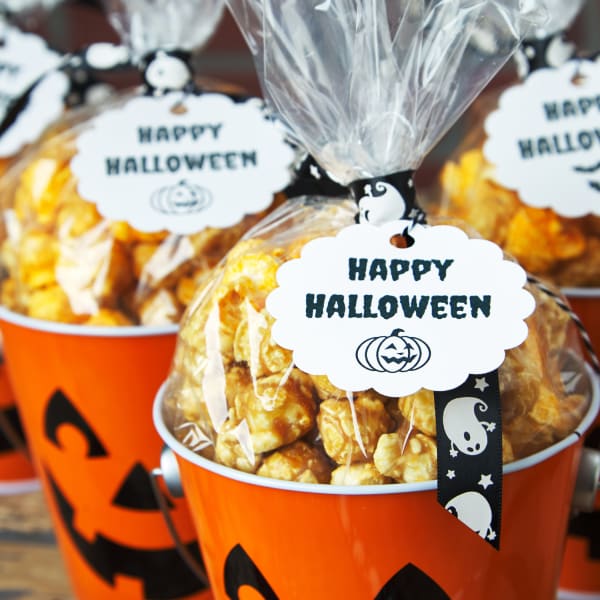 A cute "You've Been Booed" treat idea featuring orange jack o' lantern buckets and caramel corn bags. The image shows a close up view of a black and white ghost-print ribbon and Avery tag 80511 that reads "Happy Halloween."