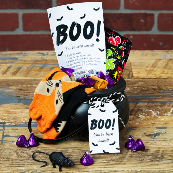 A witchy You've Been Booed idea using a plastic cauldron filled with treats. There are candies, fall-themed socks, small toys and a card with instructions. The cauldron is accessorized with a black and white string and a "Boo!" tag.
