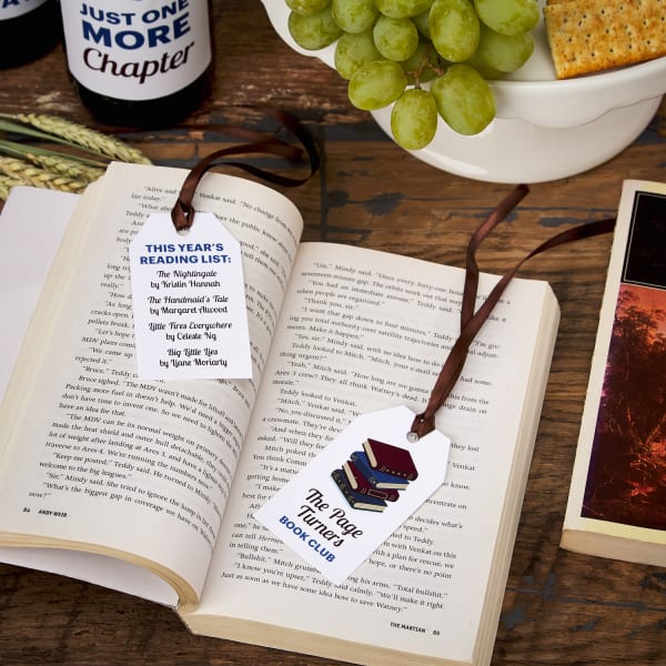 open book on table with two book club theme bookmarks made from printable tags and wine-colored ribbon next to snacks and wine bottles