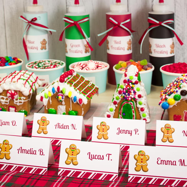 Supplies to decorate gingerbread houses and example houses are neatly lined up on a counter. In front of them are personalized gingerbread-themed tent cards for party guests. 