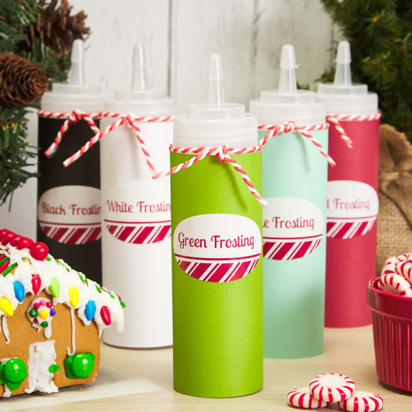 five tall, clear bottles filled with frosting in various colors and labeled with oval labels, sitting next to peppermints and a gingerbread house
