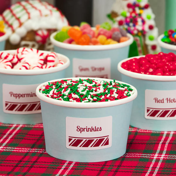 small rectangle labels with candy cane theme on light blue paper cups filled with various gingerbread house toppings on Christmas tablecloth