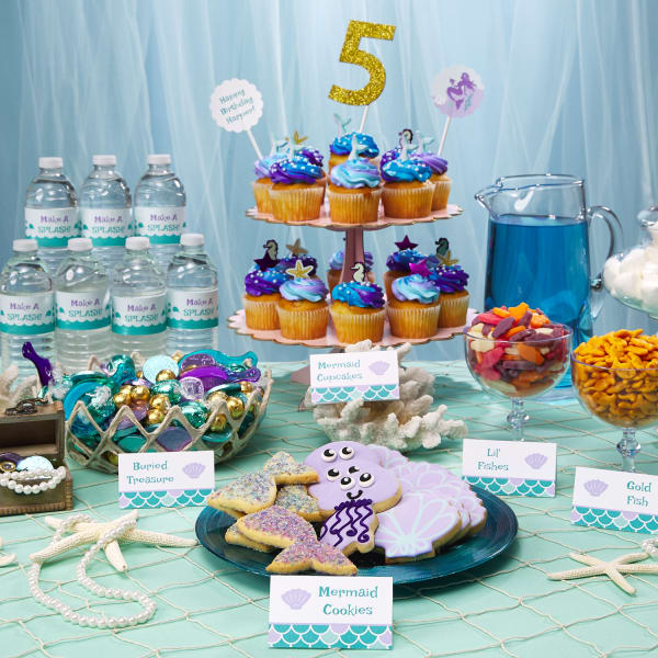 A blue and lavender party party buffet is arranged on a table. The food and drinks are personalized with Avery labels and tent cards for a Mermaid party theme.