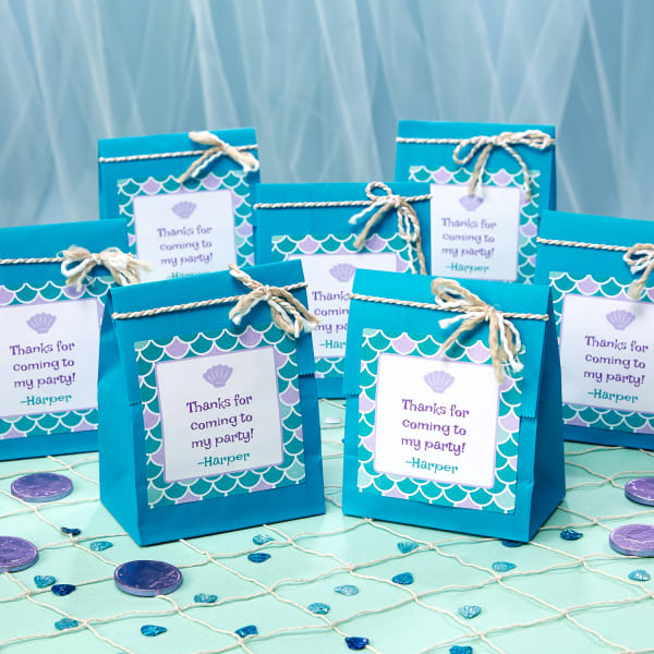 DIY party favor bags shown on an aqua table with netting and sea-themed table scatter. The bags are personalized with Avery 94100 square labels sold by the sheet.