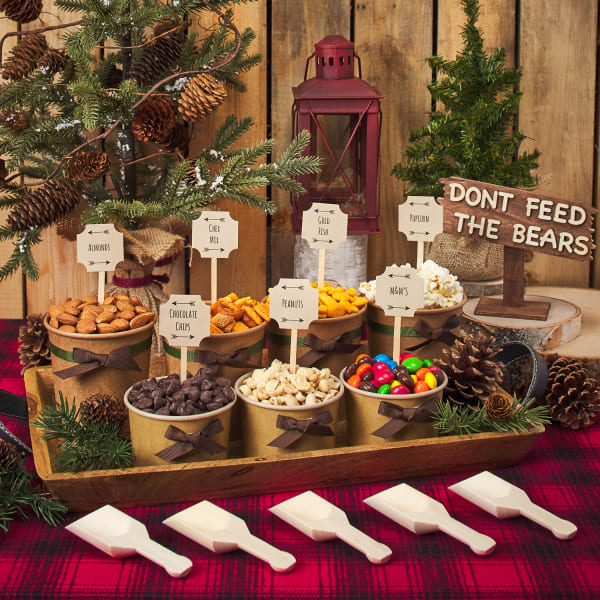 camping-themed party table with seven cups filled with snacks and labeled with scallop tags next to scoops and outdoor decor