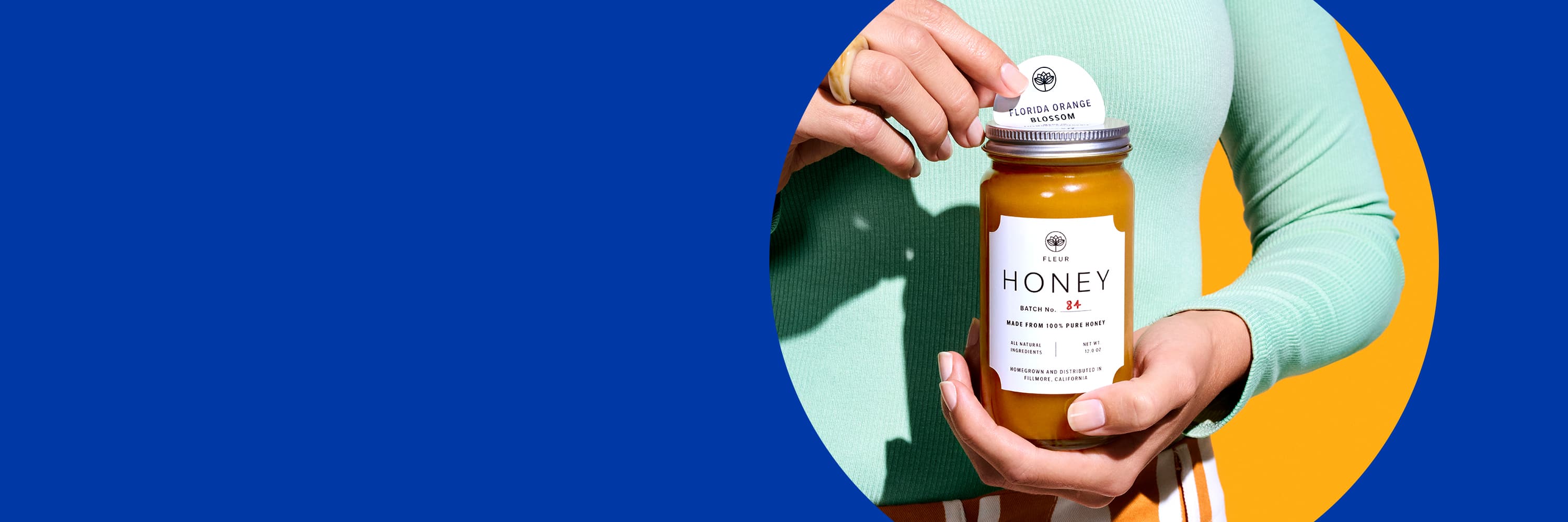 Blue banner with a photo of a person labeling the lid on a jar of honey with a custom designed round label. The person is wearing a sky blue long sleeve sweater and white and orange striped pants and is standing against a bright orange background.