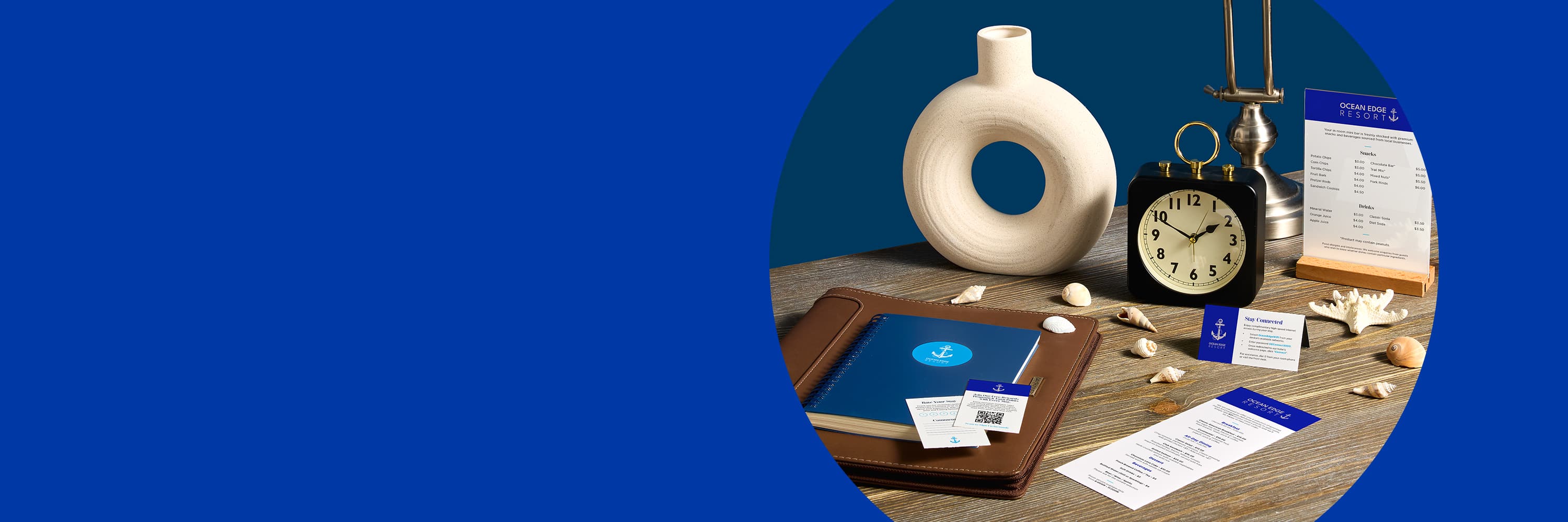 An image of a hotel room with a deep blue wall color and beach-themed decor, focused in on a wooden dresser with hotel welcome marketing materials, such as menus, tent cards, and business cards. The hotel marketing materials feature a clean deep blue and white design with anchors 