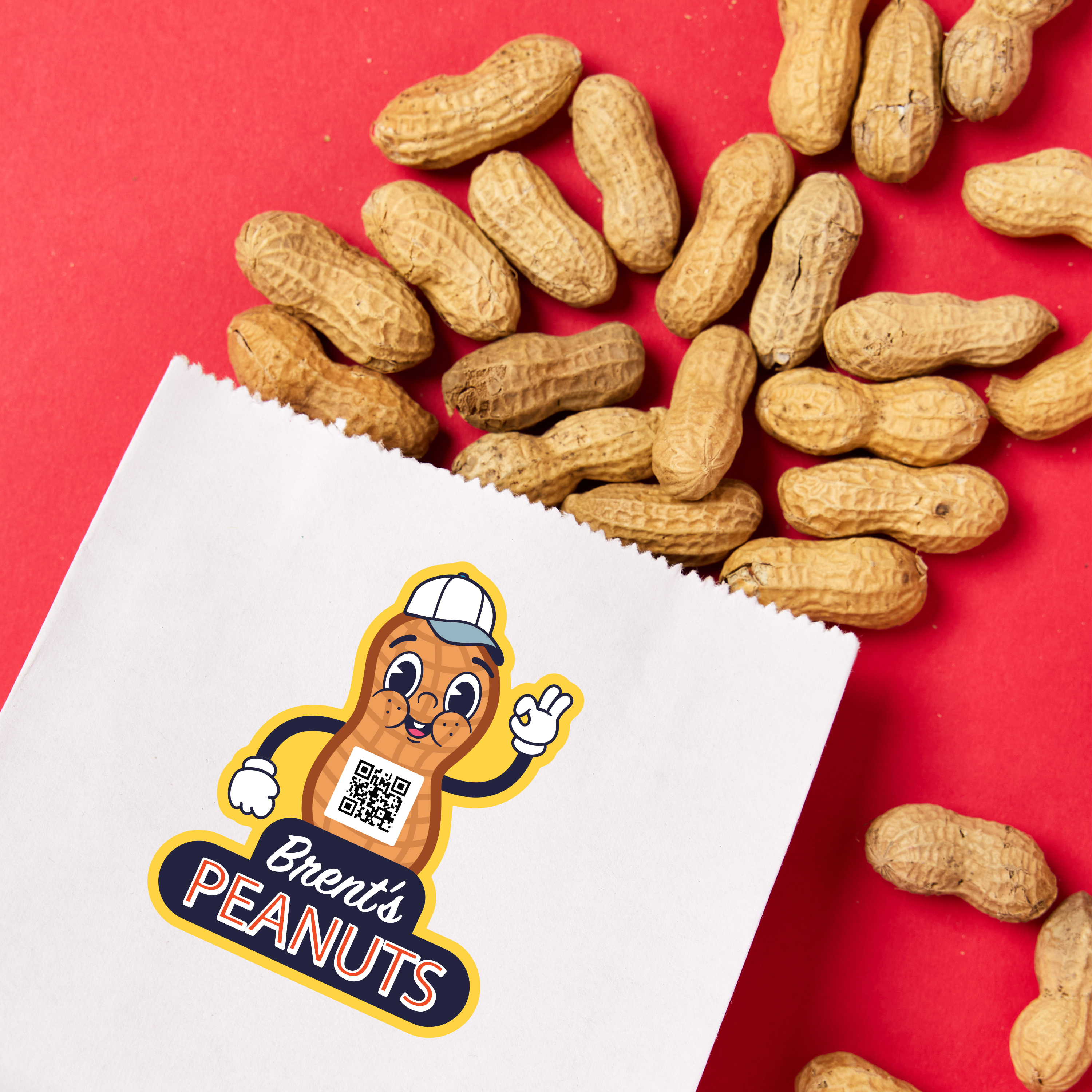A white paper bag with a custom die-cut sticker featuring a cartoon peanut mascot wearing a hard hat, white gloves, and a friendly smile. The sticker includes the text "Brent's PEANUTS" in bold blue and orange lettering, along with a small QR code on the peanut character’s chest. The bag is placed on a bright red background, with unshelled peanuts spilling out around it, creating a visually appealing contrast.