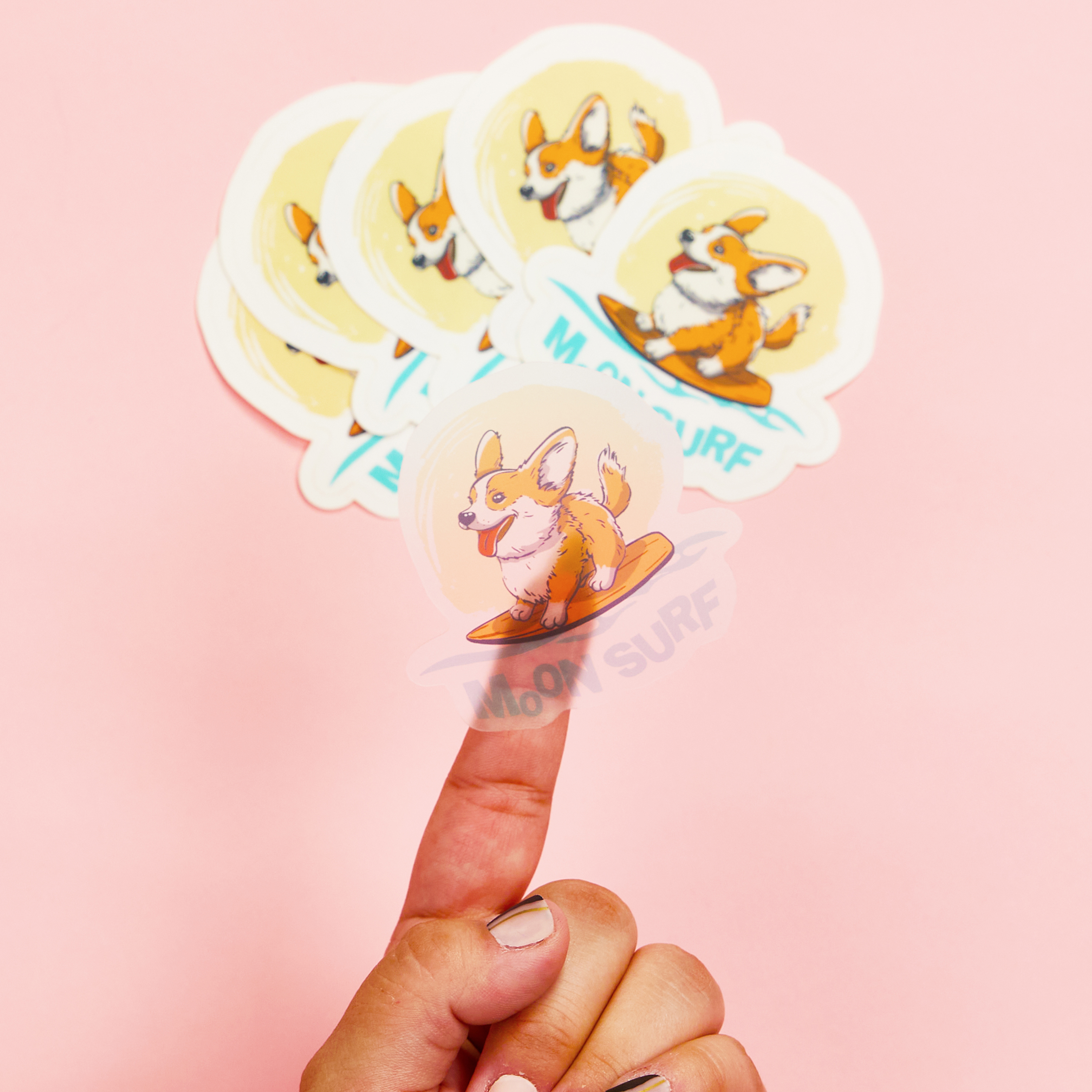 A hand holds a transparent die-cut sticker featuring an illustrated corgi surfing on a small orange surfboard. The sticker is placed on the tip of the index finger, showing its flexible, clear material. In the background, four more identical stickers with a white border and the words "MO SURF" in bold blue text are fanned out against a soft pink backdrop. The die-cut edges follow the shape of the corgi illustration, making the stickers visually distinct.