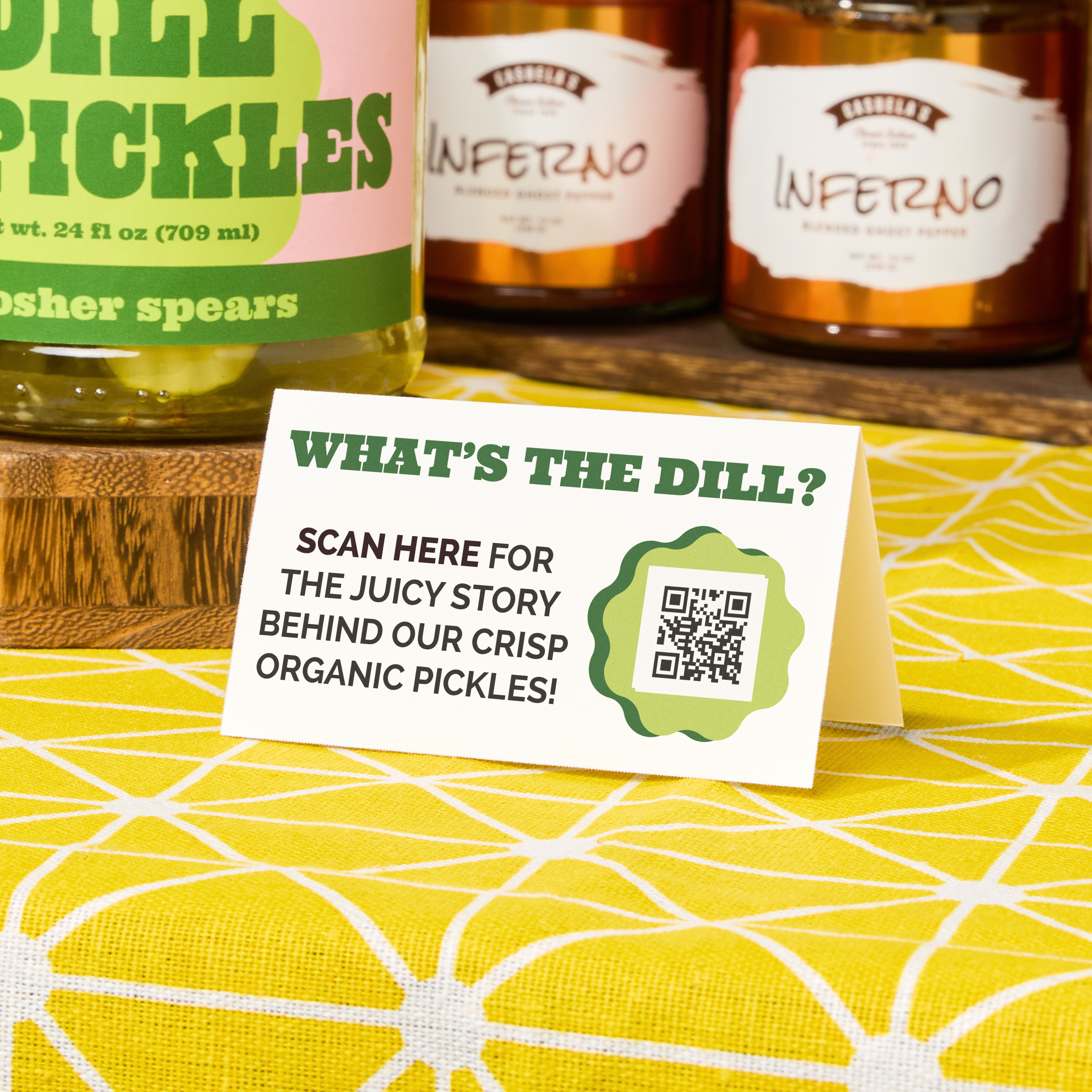 Tent card on a pickle jar display reads “What’s the Dill?” with a QR code linking to a brand story, shown on a yellow patterned tablecloth.