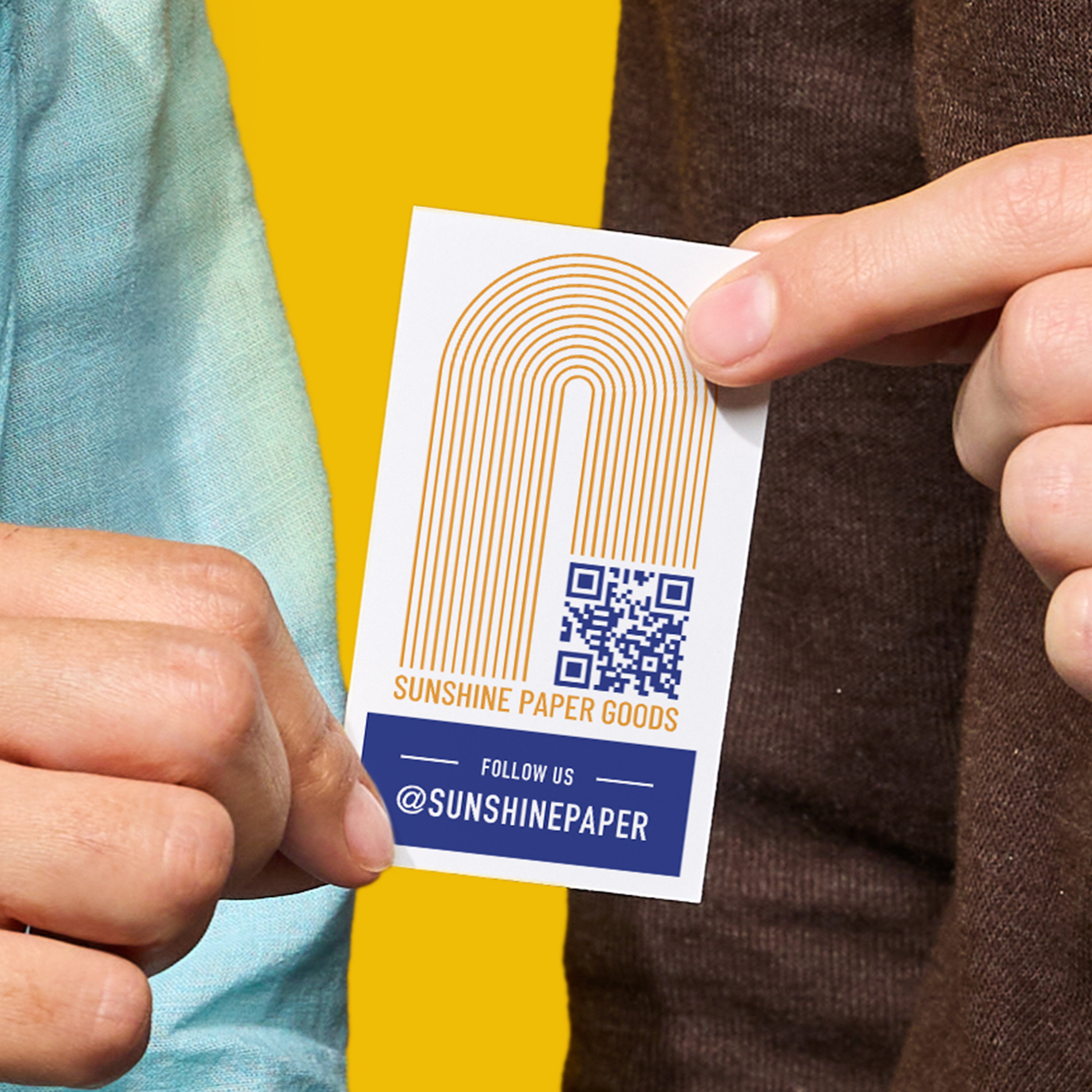 Close-up of two hands exchanging a business card that features a logo, QR code, and social media handle against a bold yellow backdrop.
