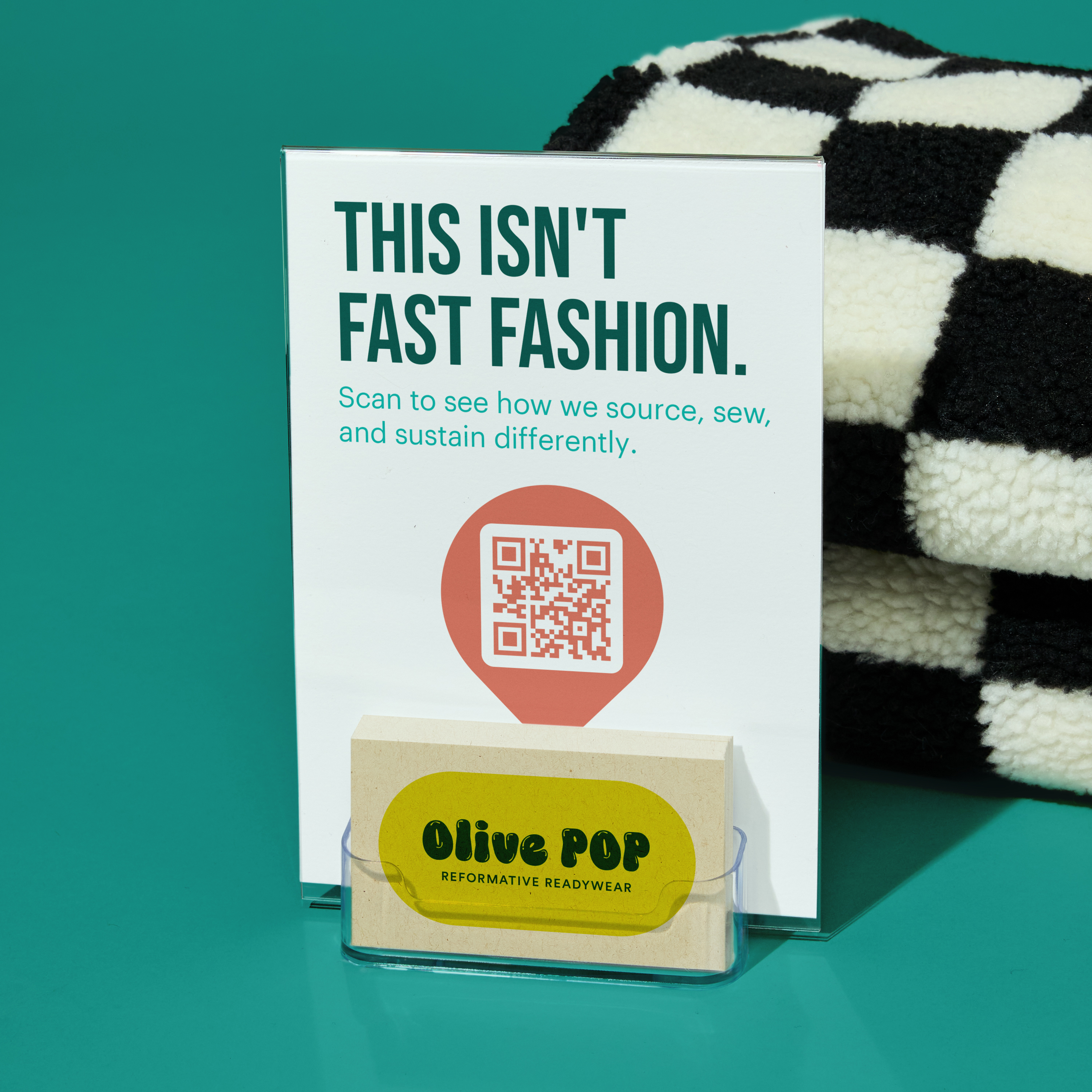 A tabletop business card display with a standup sign reading “This isn’t fast fashion” and an attached business card holder filled with cards for “Olive Pop.” The display sits in front of a folded checkered textile on a teal surface.