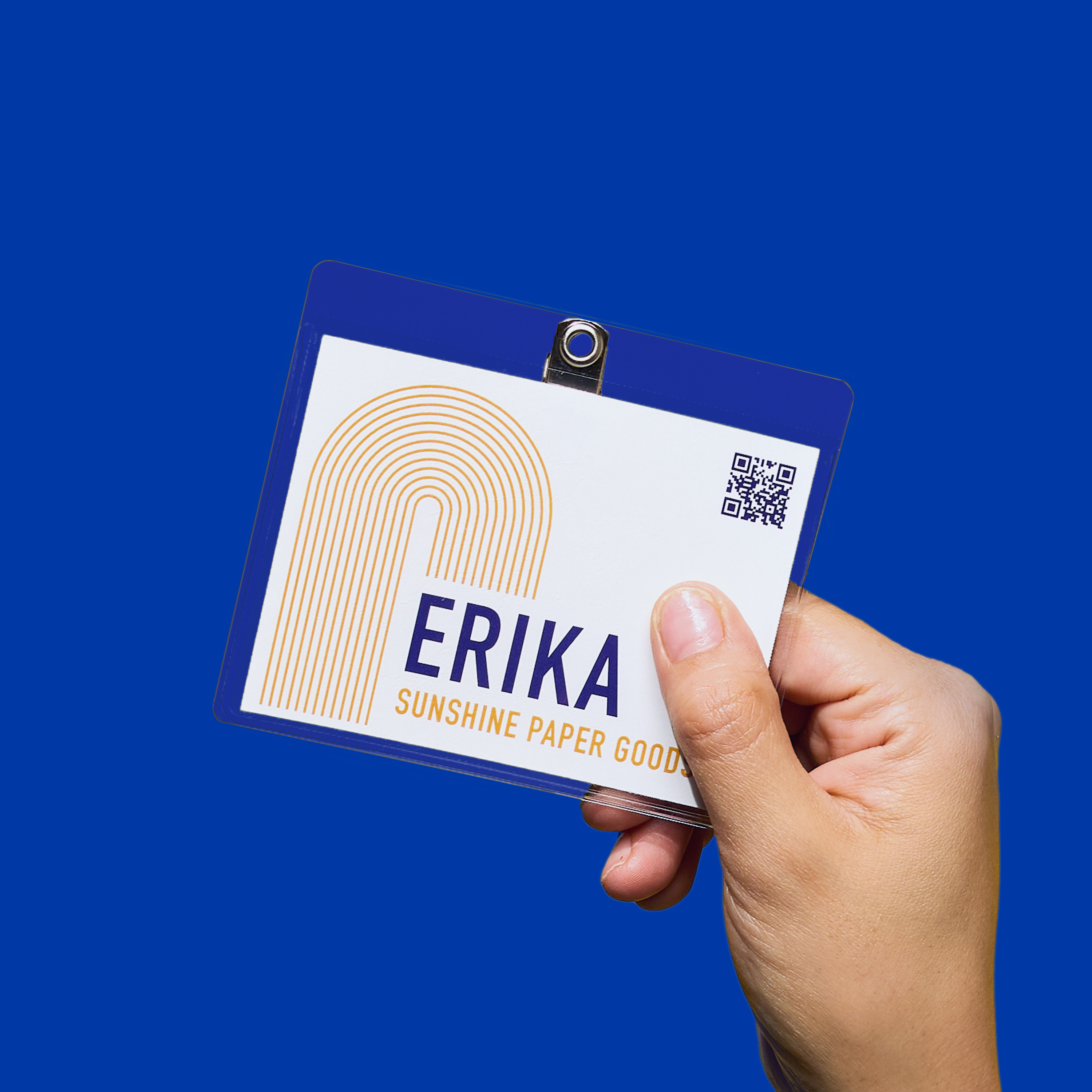 A hand holds a clip-style name tag labeled “Erika – Sunshine Paper Goods” against a vibrant blue background, showing personalized expo branding.