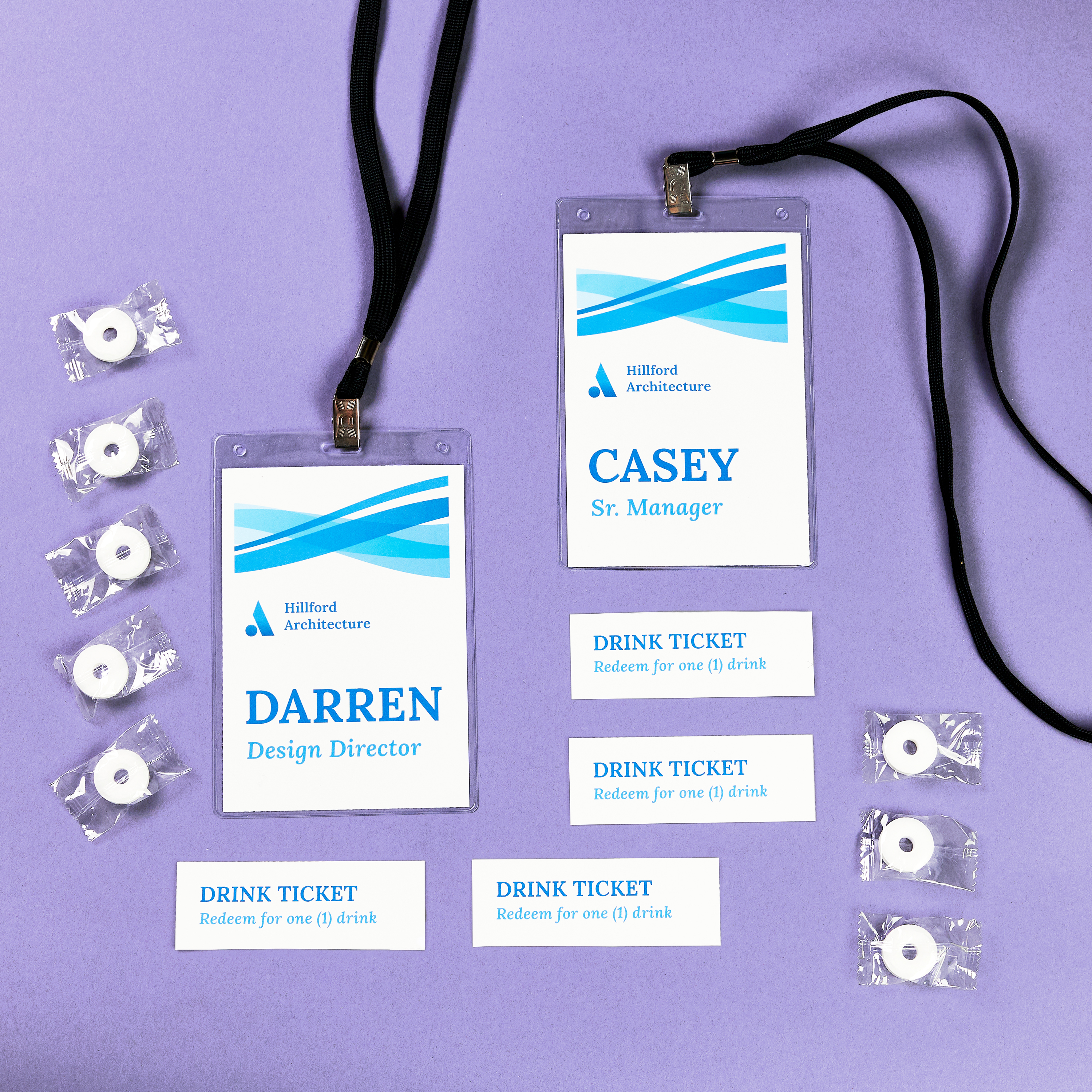 Two vertical name badges labeled “Darren” and “Casey” with lanyards, surrounded by clips and individually wrapped mints, arranged on a lavender background for a trade show setup.