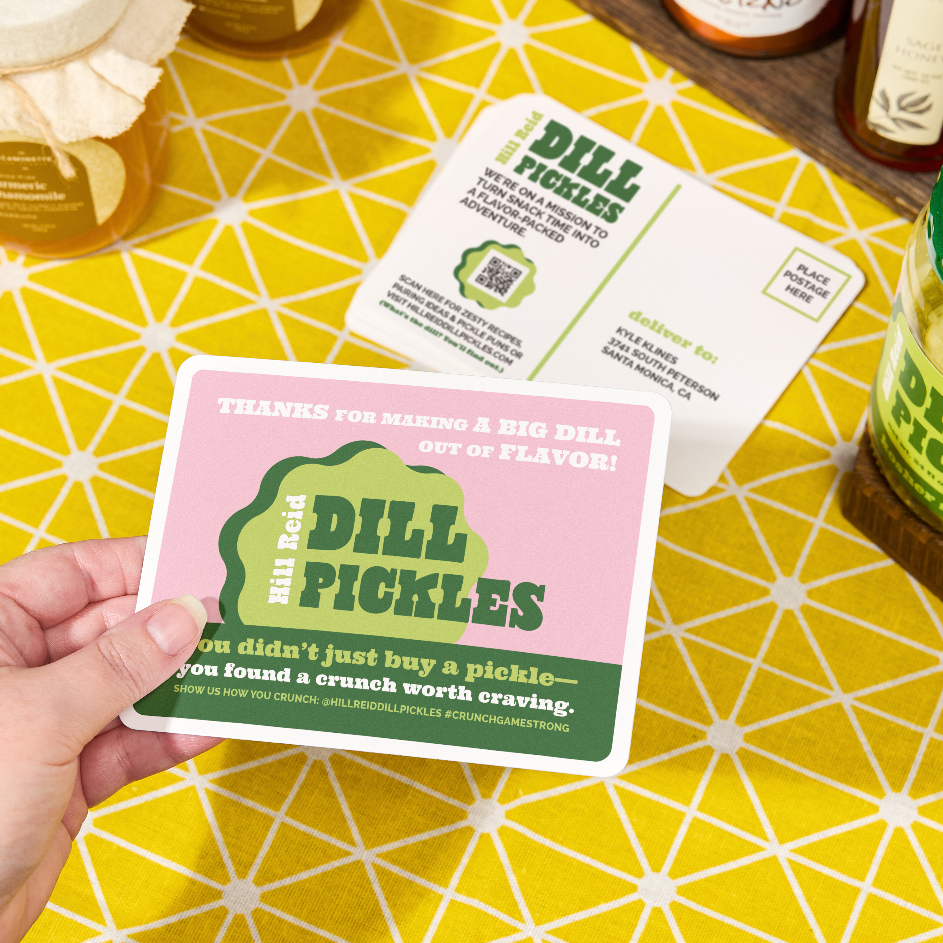 A hand holds a pink postcard with a QR code and bold “Dill Pickles” branding, shown on a yellow patterned table alongside a matching mailer and glass jars.