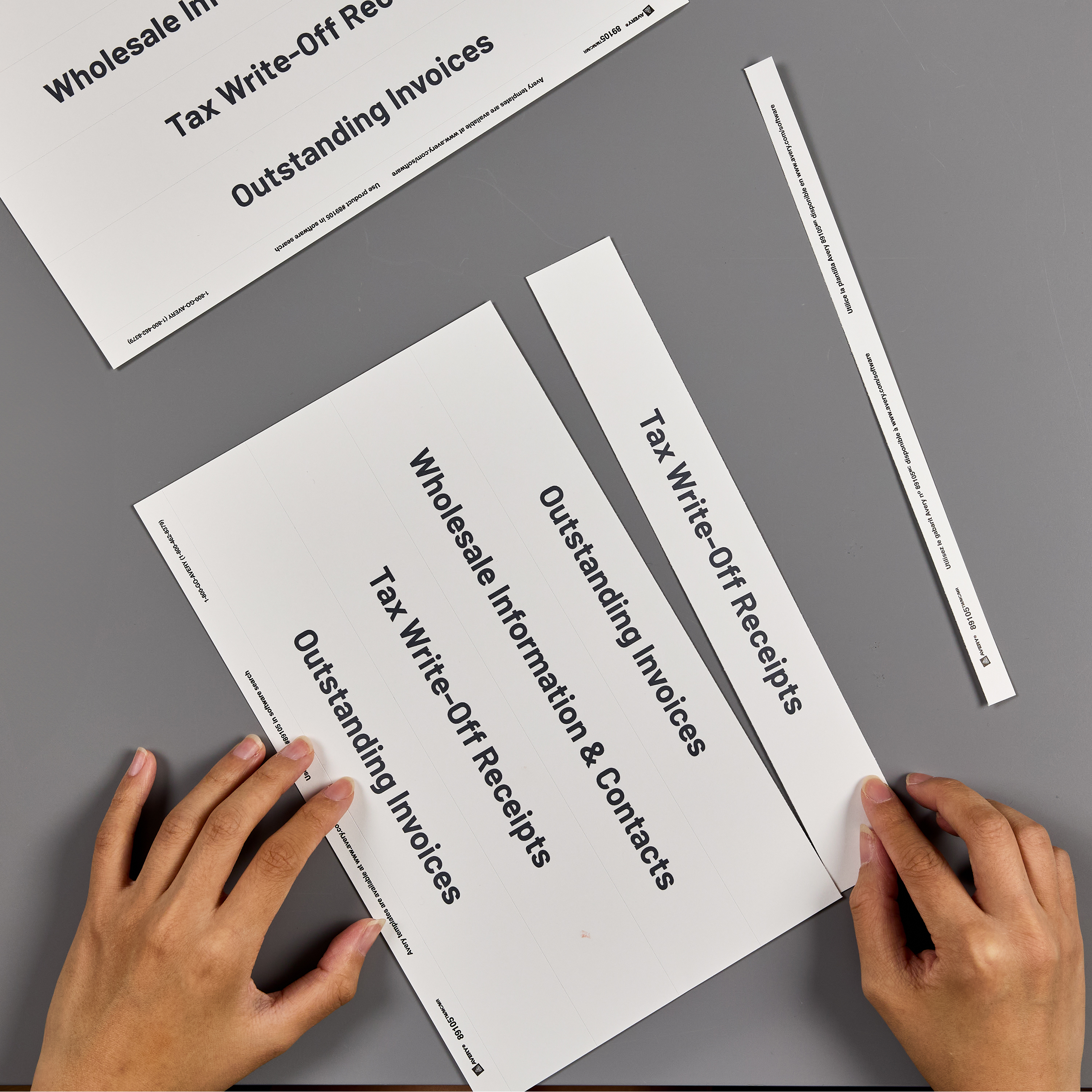 Hands placing a custom binder spine insert labeled 'Tax Write-Off Receipts' on a gray surface, with additional spine inserts for 'Wholesale Information & Contacts' and 'Outstanding Invoices' nearby.