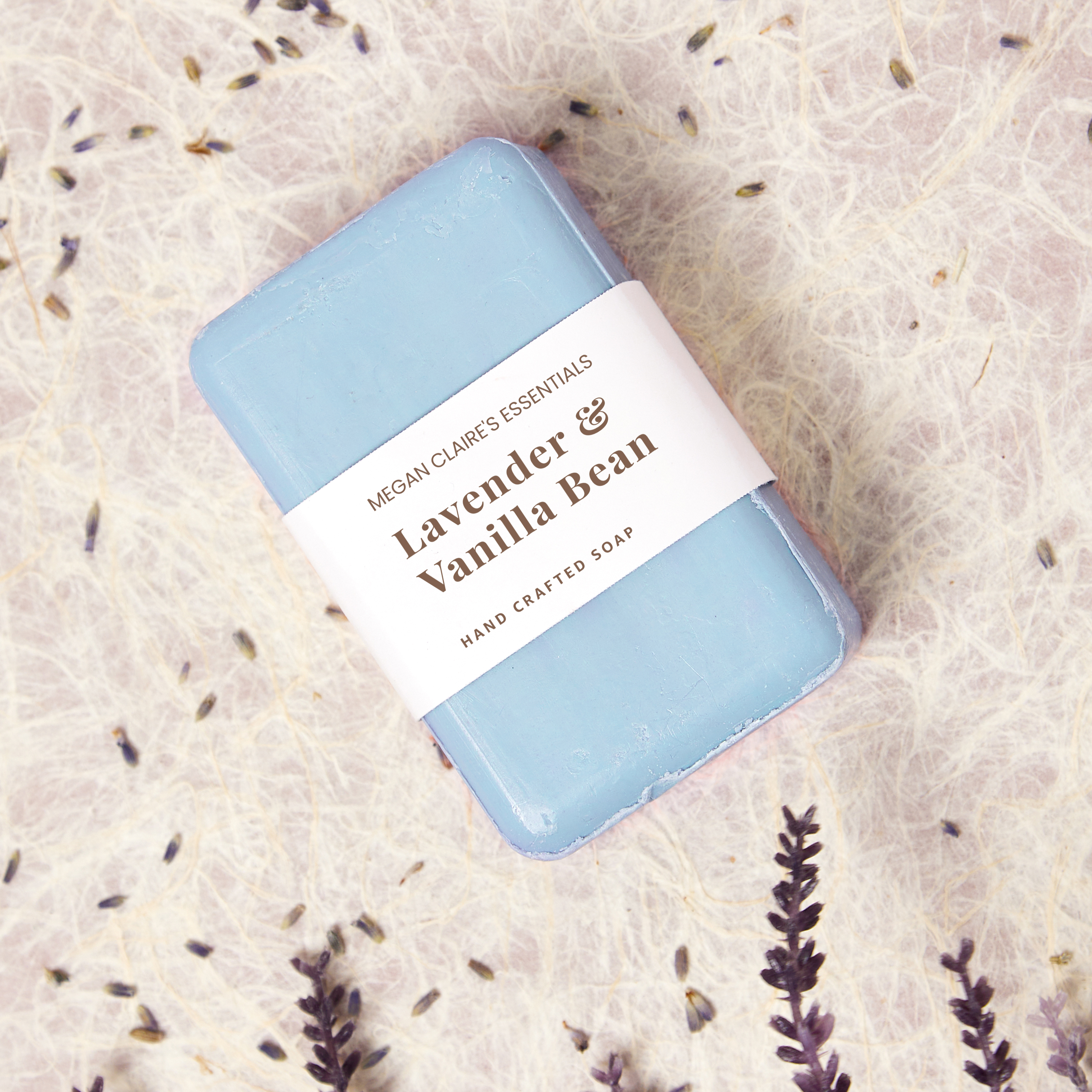 Blue handmade soap bar wrapped with a minimalist white paper band labeled 'Lavender & Vanilla Bean,' designed using an Avery Binder Spine Insert 89105, surrounded by dried lavender on a textured, natural background.