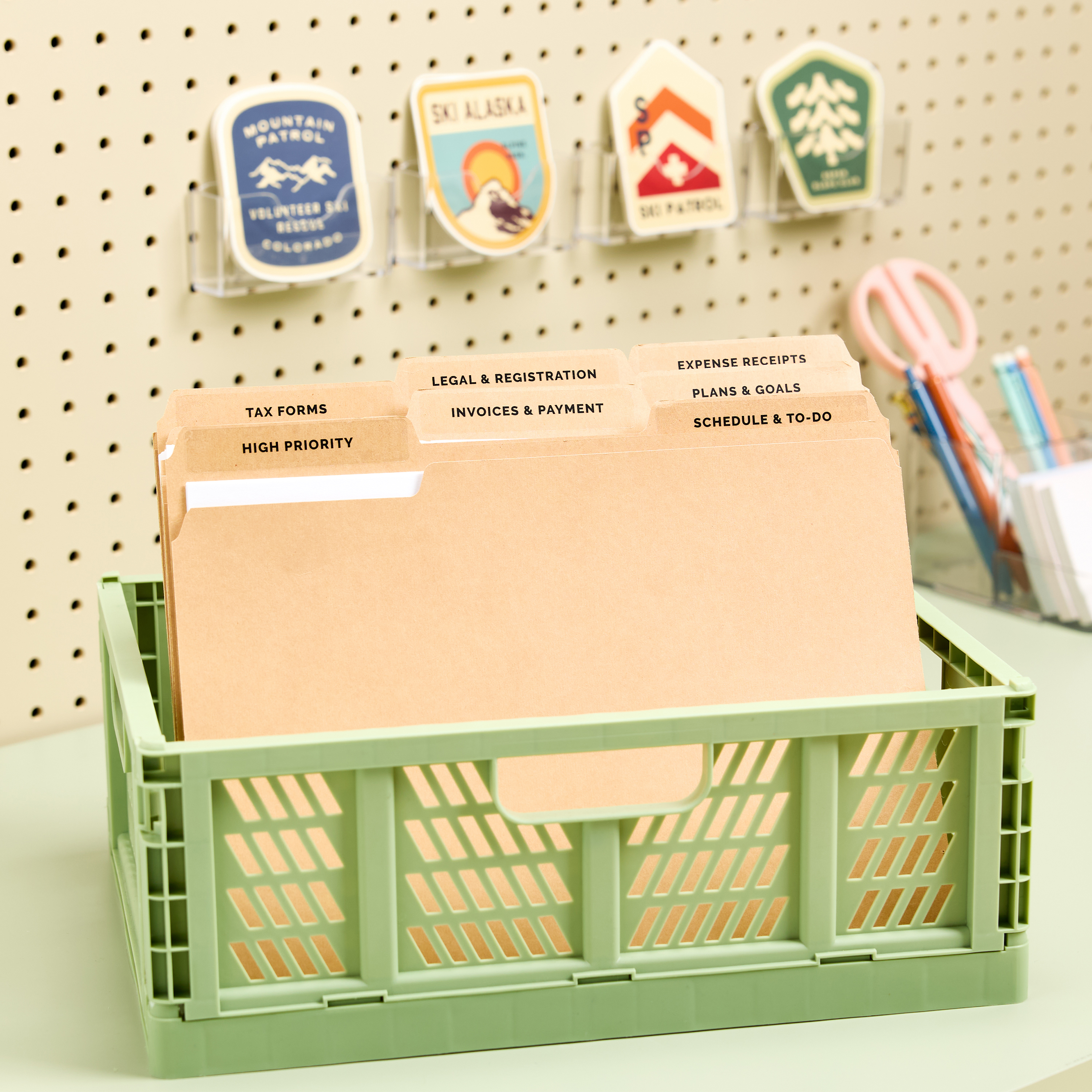 A mint green storage crate holds file folders neatly labeled with clear Avery 5029 file folder labels. The labels include categories like "Tax Forms," "Invoices & Payment," and "Schedule & To-Do," demonstrating a desk organization idea to sort and store active paperwork with a system. The crate is placed on a tidy workspace with additional office supplies and decorative wall accents in the background.