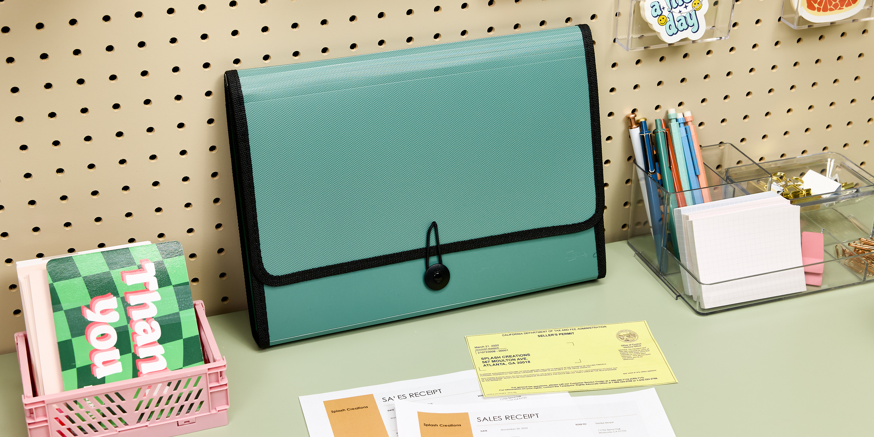 A sage green Avery 73555 file organizer is displayed on a desk, neatly holding important documents like sales receipts and permits. The workspace features additional desk organization ideas, including a pink basket with thank-you cards, a clear organizer with pens, paper clips, and sticky notes. The pegboard wall in the background adds functional storage. This setup demonstrates practical tips on how to organize your desk for efficiency and tidiness.