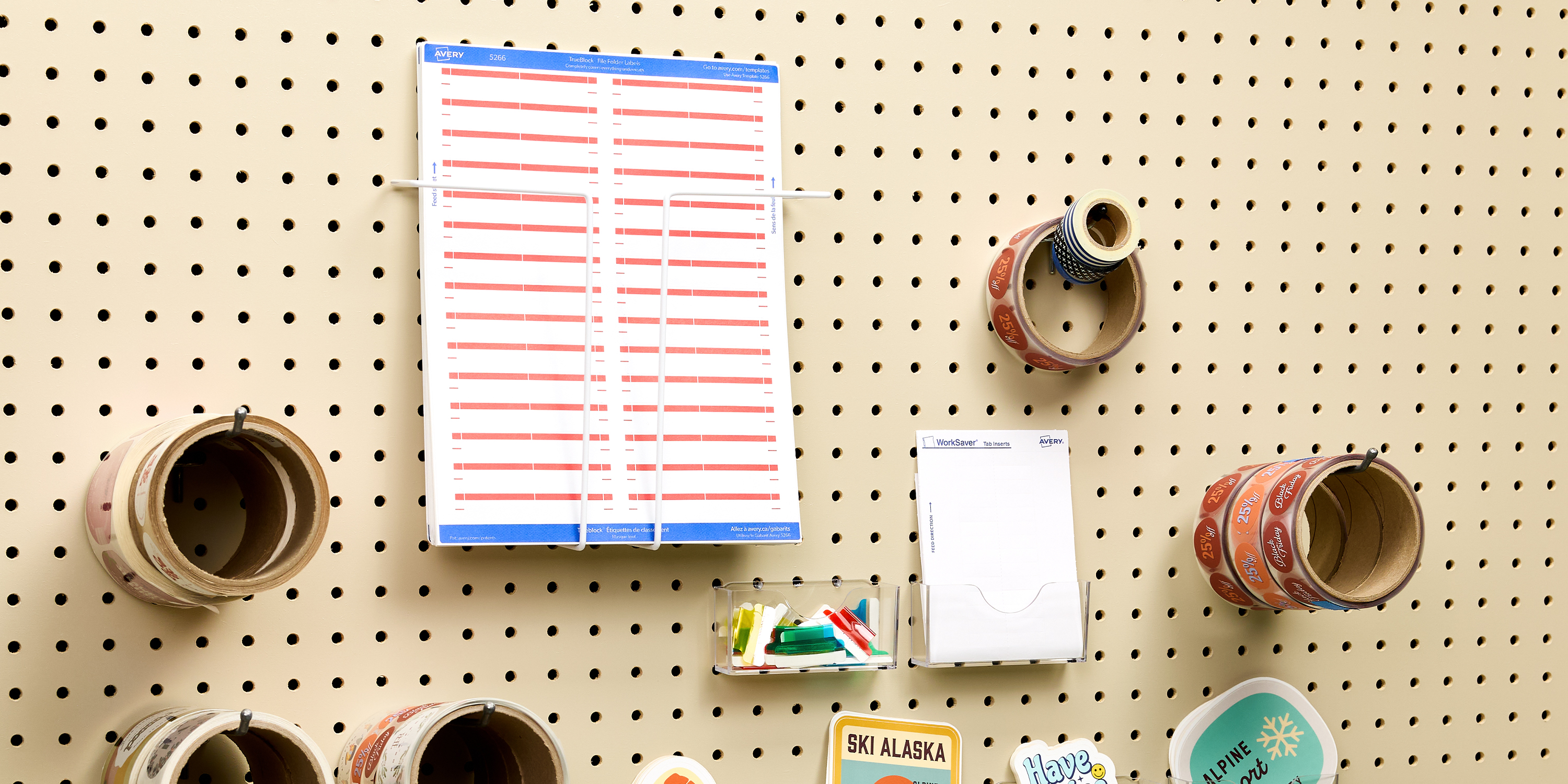 A pegboard wall demonstrates how to organize a desk using vertical space. The setup includes rolls of washi tape neatly hung on hooks, a clipboard-style schedule, a notepad holder, and a clear compartment containing push pins and small clips. Decorative elements like travel-themed patches add personality while keeping the area functional and tidy.