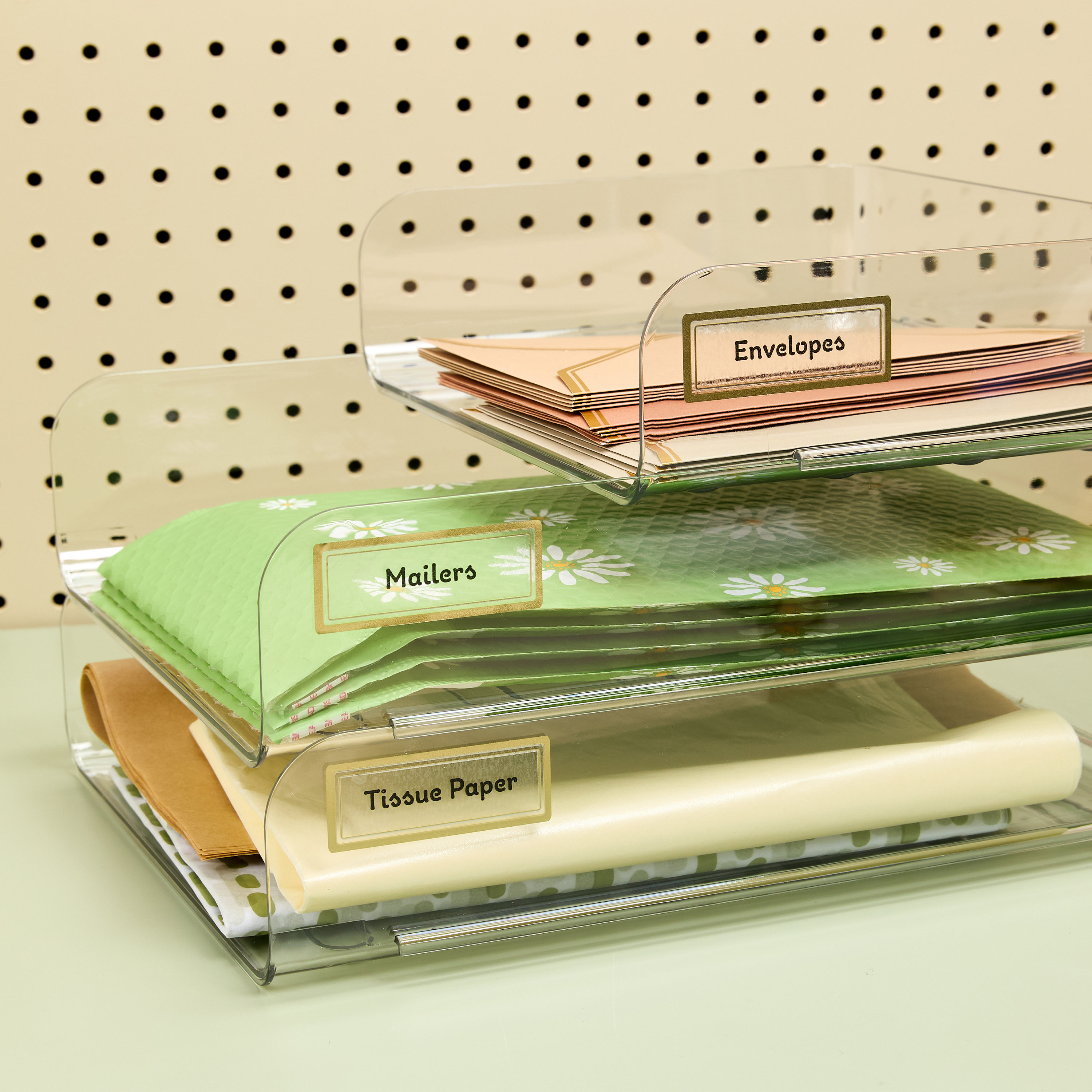 A set of clear stacking trays organizes envelopes, mailers, and tissue paper, each labeled with Avery 6550 clear address labels featuring a gold border. The labels add a stylish and practical touch to the workspace, making it easy to find supplies. This setup demonstrates a simple, yet stylish, solution for how to organize your desk and keep it tidy.