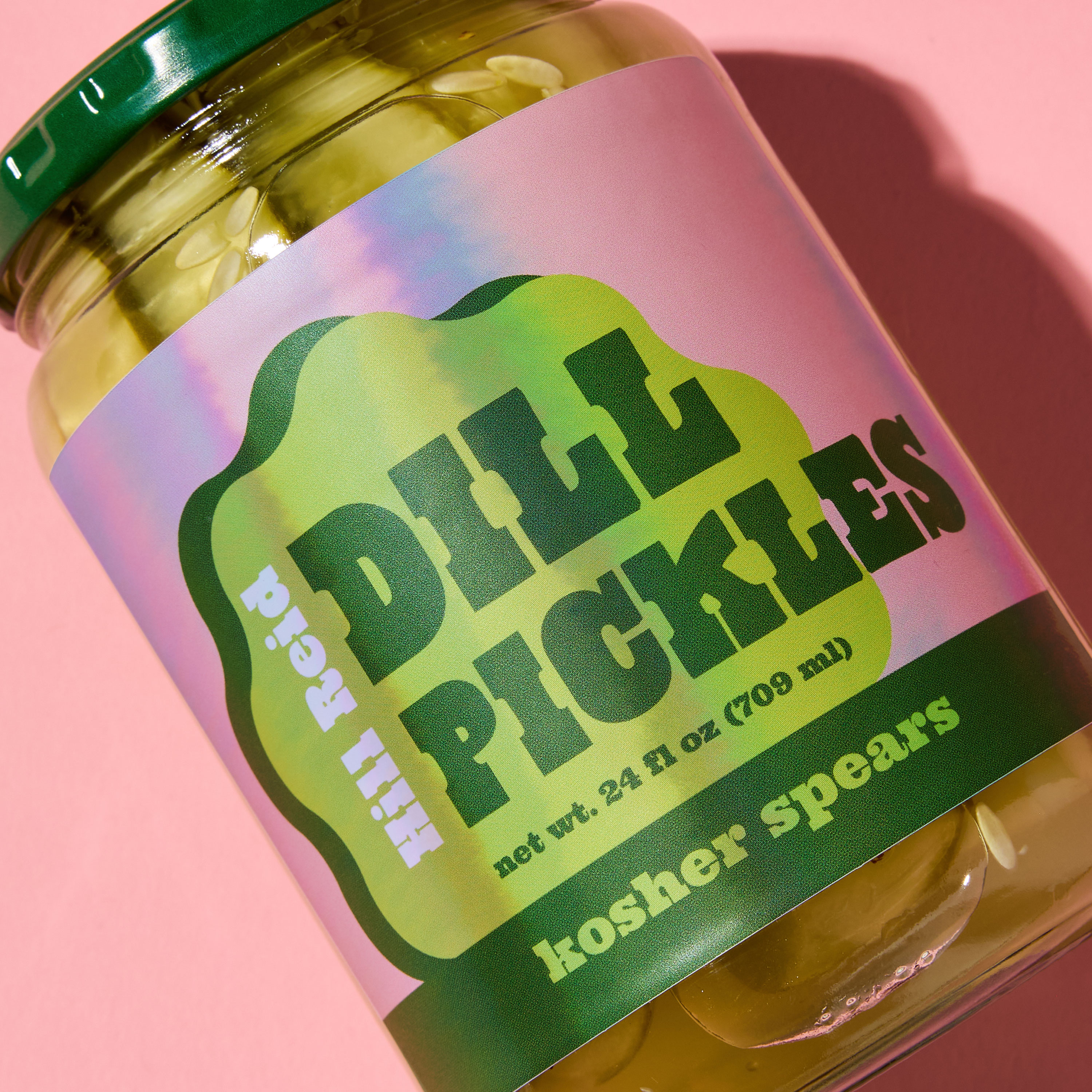 A close up of a pickle jar on a pink background. The pickles are labeled with a rainbow holographic film that reads "Hill Reid Dill Pickles." The label design is pink and green, but the holographic film gives shades of blues and purples where the light hits.
